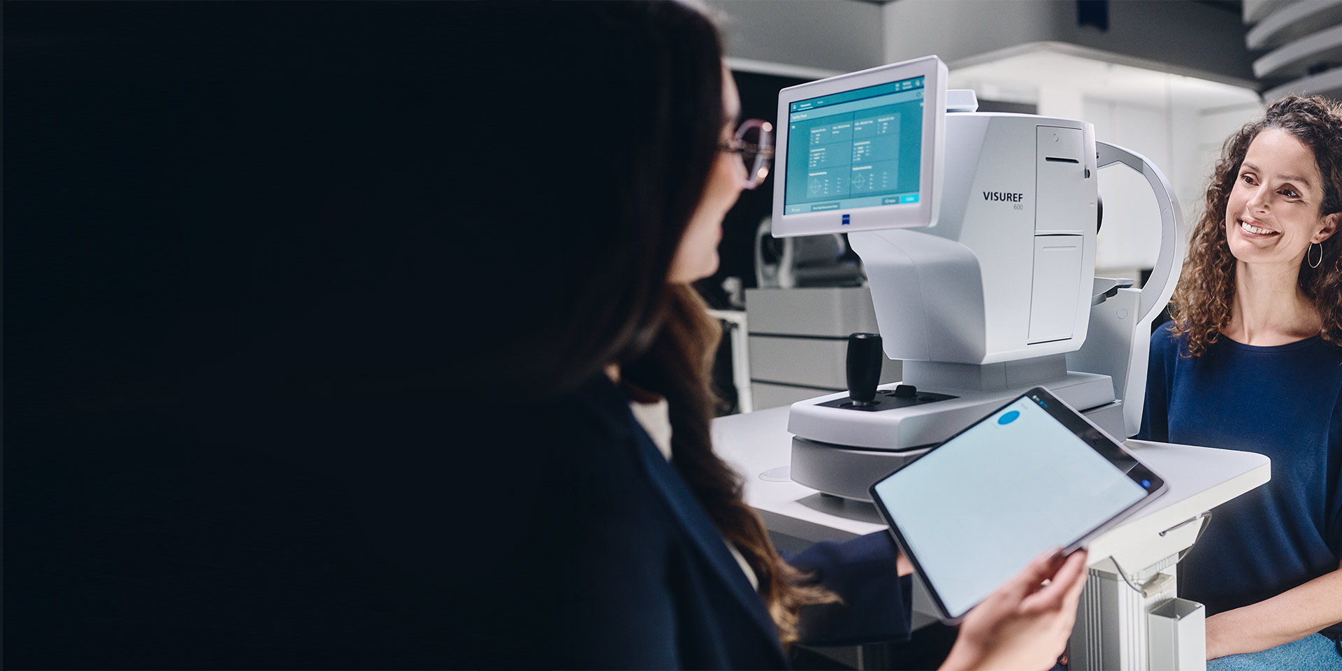 Optička sedící před přístrojem ZEISS VISUREF 600, dívající se na svůj tablet a hovořící se zákazníkem.