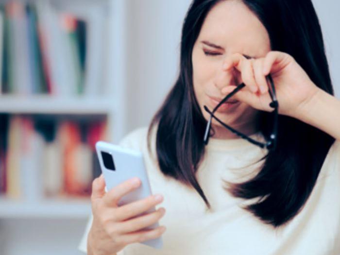 Obrázek zobrazující 30letou ženu, která se dívá na svůj telefon a protírá si přitom oči.