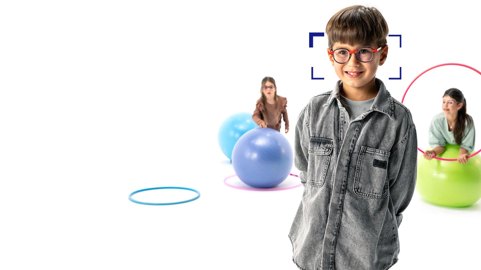 Hnědovlasý chlapec s kulatými brýlemi osazenými brýlovými čočkami ZEISS MyoCare stojí v popředí a usmívá se do objektivu. Na pozadí jsou dvě dívky, které mají nasazené brýlové čočky ZEISS MyoCare a hrají si s obručemi a gymnastickými balóny.