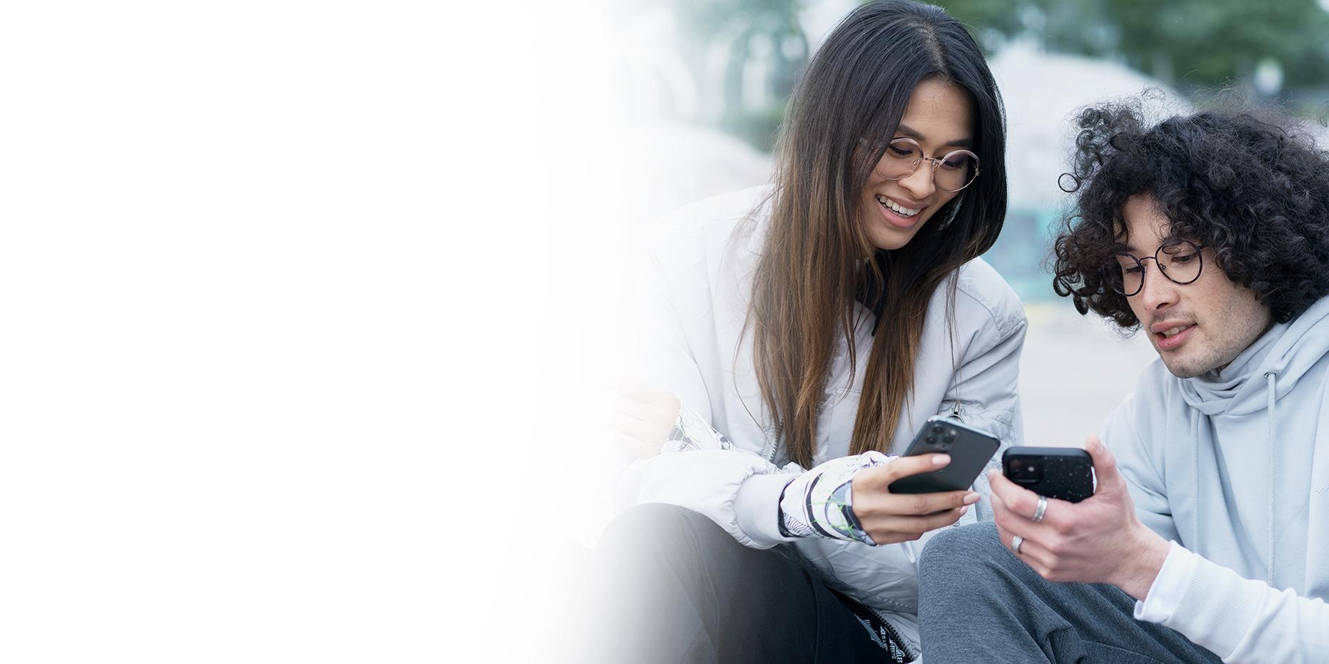 Dva mladí lidé, usmívající se žena s dlouhými hnědými vlasy a chlapec s kudrnatými černými vlasy, se v brýlích ZEISS SmartLife dívají do svých telefonů. 