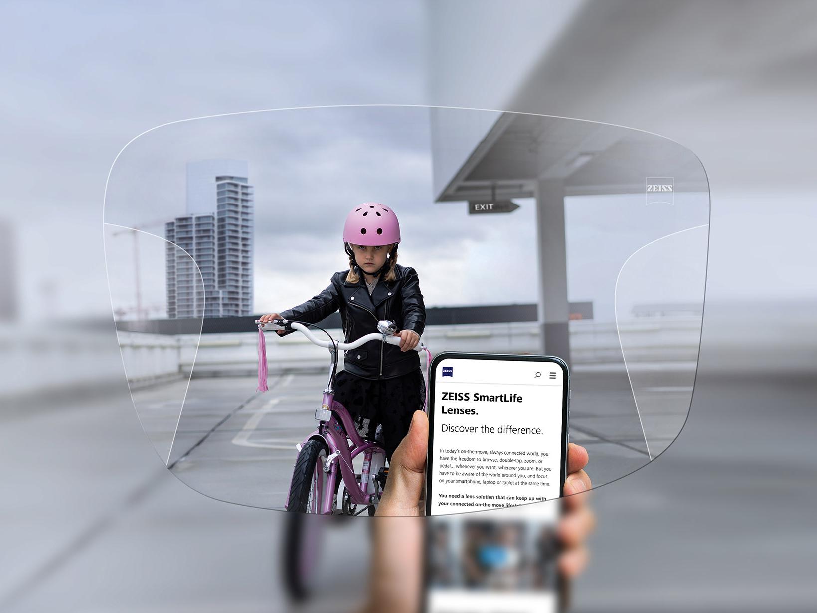Osoba dívající se na svůj mobil přes progresivní brýlové čočky ZEISS SmartLife. Na pozadí je dívka na kole v růžové přilbě. 
