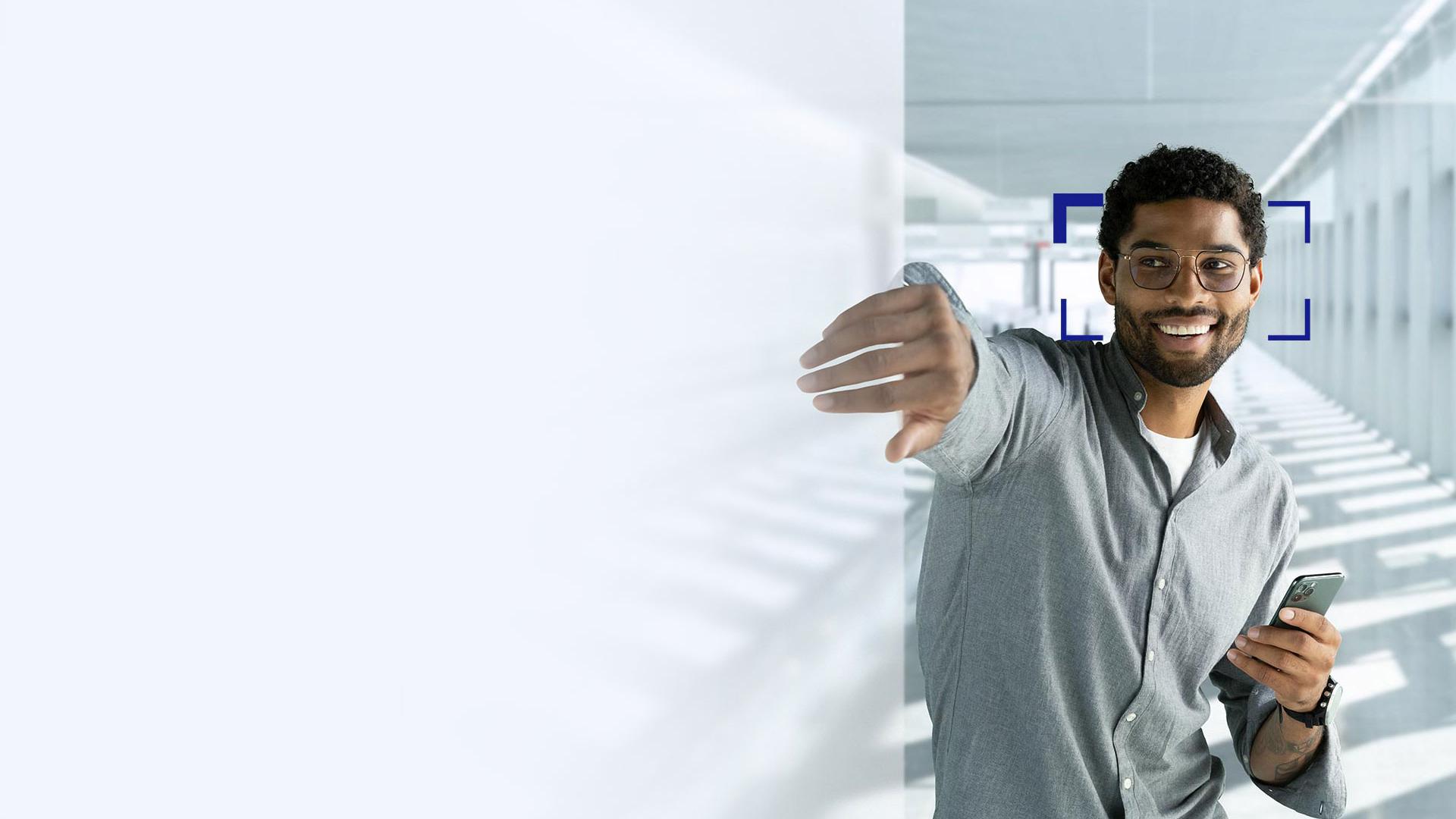 Mladý muž s černými vlasy a nasazenými brýlemi s brýlovými čočkami ZEISS SmartLife se usmívá a natahuje ruku dovnitř obrazovky.