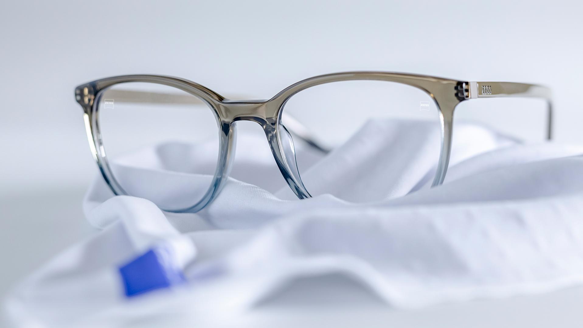 Brýle s šedomodrými obrubami a čočkami ZEISS opatřenými vrstvou DuraVision® leží na bílém hadříku z mikrovlákna.