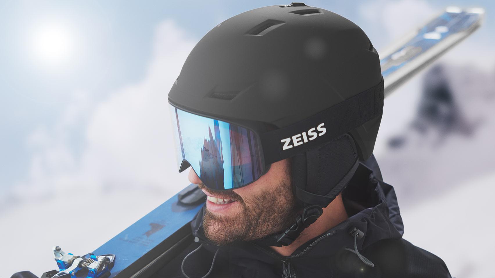 Muž v černé přilbě s modrými zrcadlovými lyžařskými brýlemi ZEISS s lyžemi opřenými o pravé rameno.