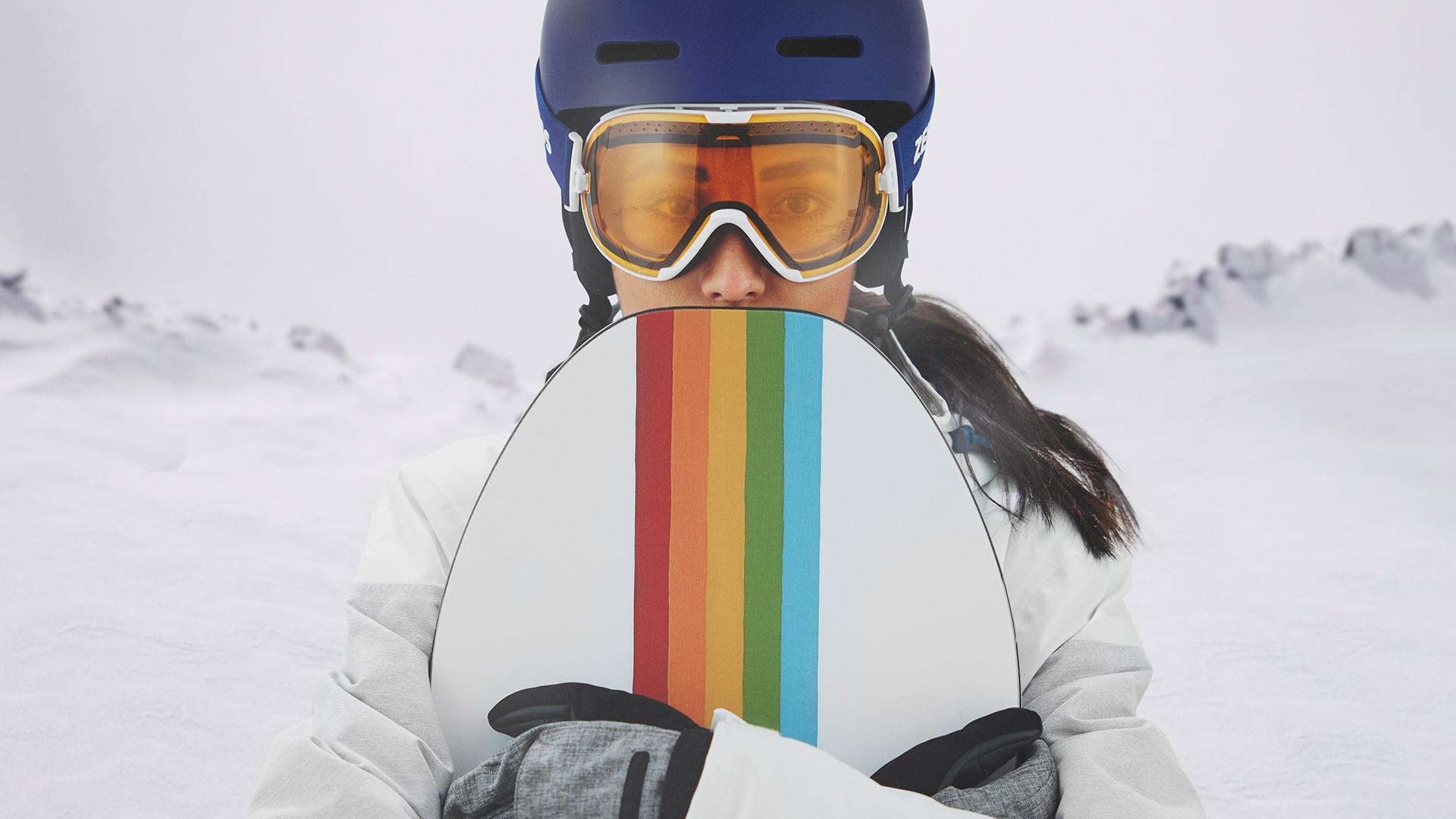 Mladá žena v lyžařských brýlích opatřených technologií výměny skel ZEISS Interchangeable, která v ruce drží snowboard.