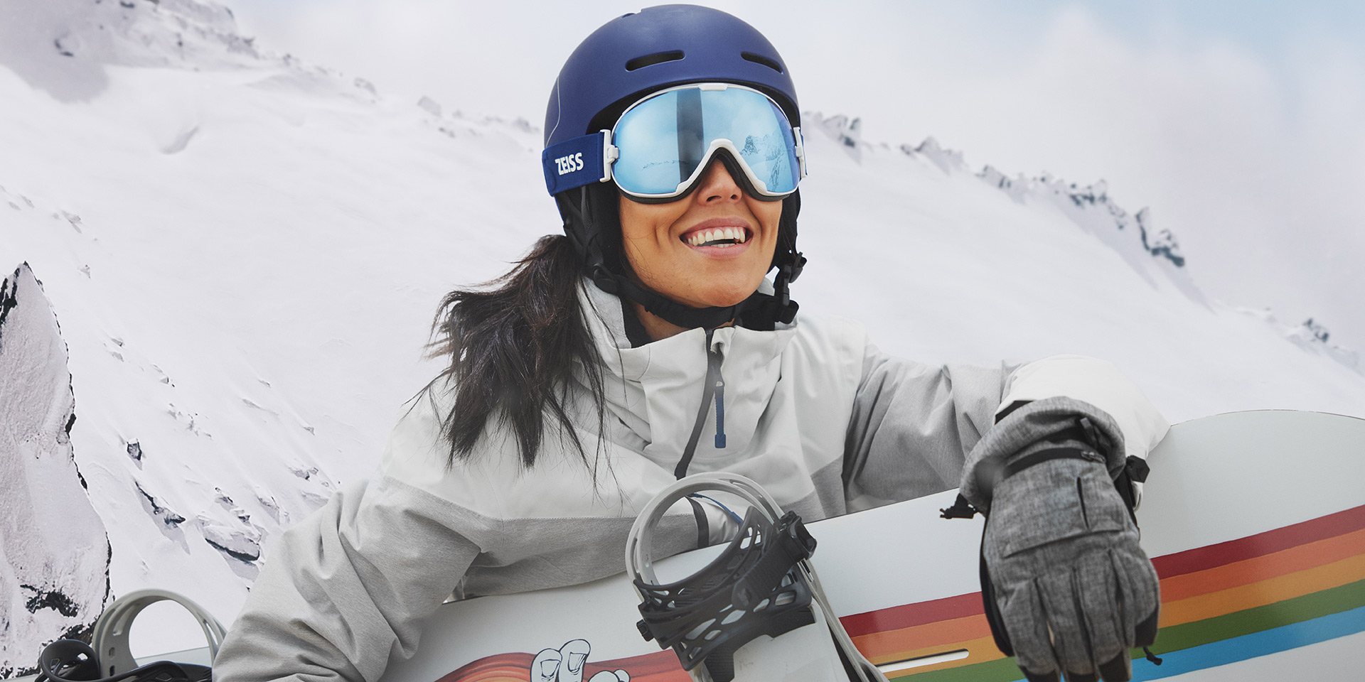 Mladá žena v lyžařských brýlích opatřených technologií výměny skel ZEISS Interchangeable, která v ruce drží snowboard.